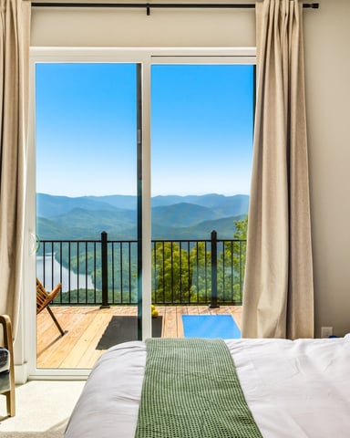 Bedroom with bed, black leather chair, and glass doors opening to balcony overlooking mountains and river valley