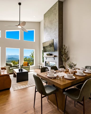 Open-plan living and dining room with mountain views through large windows, leather sofa, wood dining table, and ceiling fan.