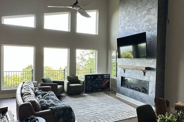 Modern living room with dark wood floors, black furniture, ceiling fan, and floor-to-ceiling windows overlooking landscape