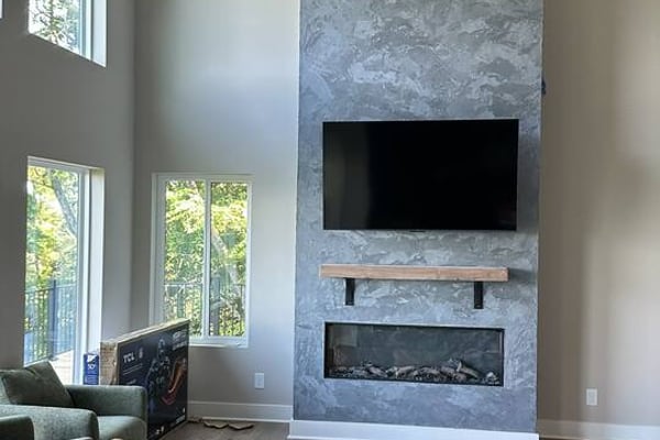 Living room with concrete fireplace wall, mounted TV, linear fireplace insert, ceiling fan, brown leather sofa, and tall windows with forest view